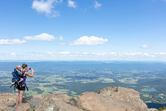 A woman with a baby on her back takes a picture with her phone on top of a mountain summit.