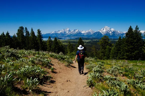 A hiker walks down a trail towards a mountain range.
